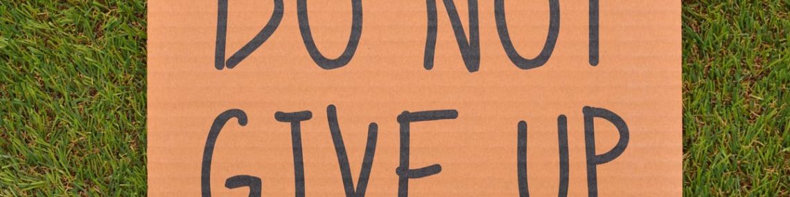 Cardboard sign reading 'Do Not Give Up' placed on green turf, symbolizing perseverance.